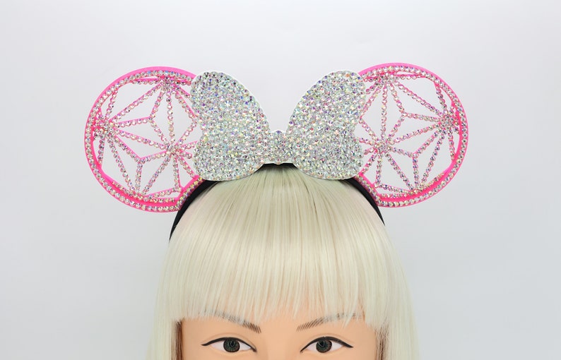 May include: Pink mouse ears headband with a large, sparkly, silver bow and a geometric design on the ears.
