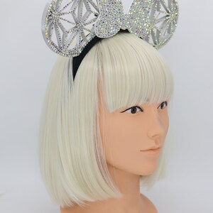 May include: A silver headband with a large, sparkly bow and two round, geometric ears. The ears are covered in rhinestones and have a white, translucent finish.