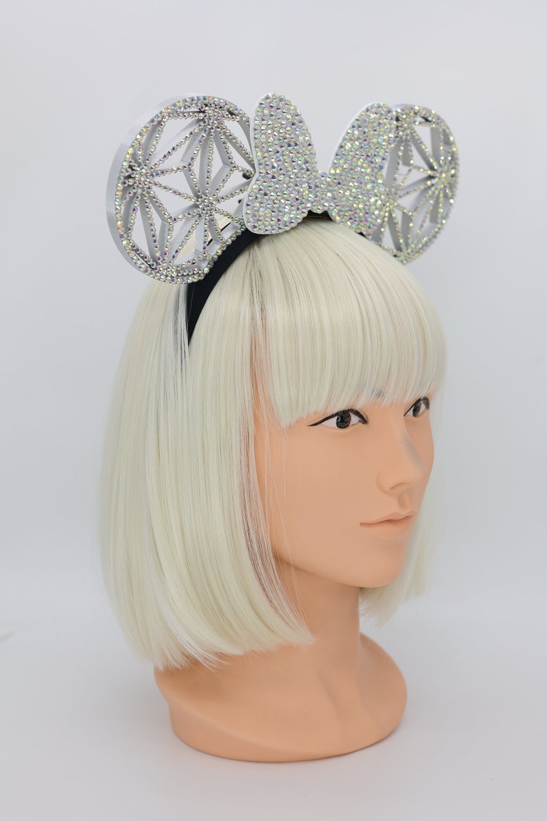 May include: A silver headband with a large, sparkly bow and two round, geometric ears. The ears are covered in rhinestones and have a white, translucent finish.