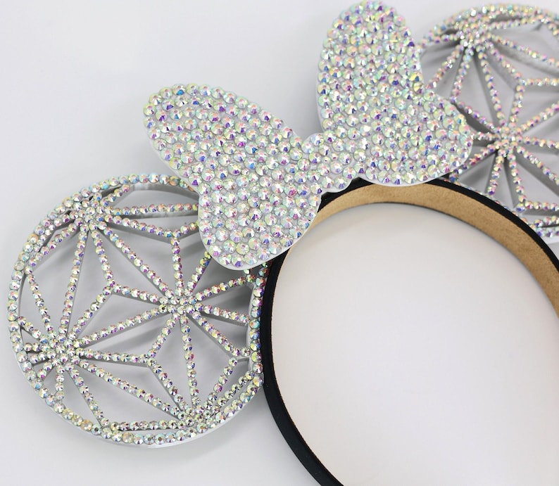 May include: A pair of silver sparkly mouse ears with a black headband. The ears are covered in iridescent rhinestones and have a geometric design.
