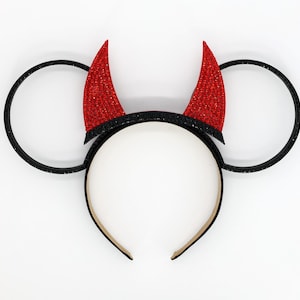 May include: A black glitter headband featuring two circular black glitter ears and two red glitter devil horns positioned between the ears. The headband has a smooth, light-colored interior lining and a dark, textured exterior finish throughout.
