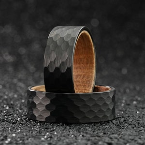 May include: Two black rings with a hammered finish. The top ring has a wooden inlay. The rings are on a black textured surface.