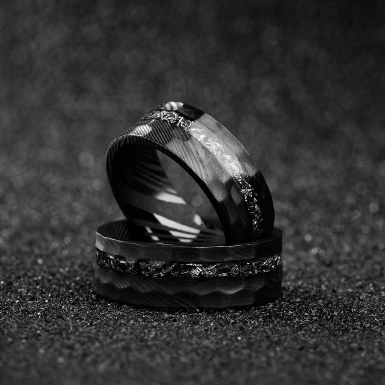 May include: Two black rings with textured surfaces are stacked on top of each other. The top ring has a patterned design and a decorative band. The bottom ring has a hammered texture and a band of small, irregular shapes.