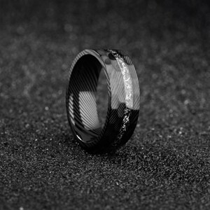 May include: A black and white ring with a patterned design. The ring has a band of lighter material with a textured surface. The ring is set against a dark, textured background.