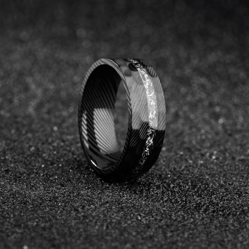 May include: A black and white ring with a patterned design. The ring has a band of lighter material with a textured surface. The ring is set against a dark, textured background.