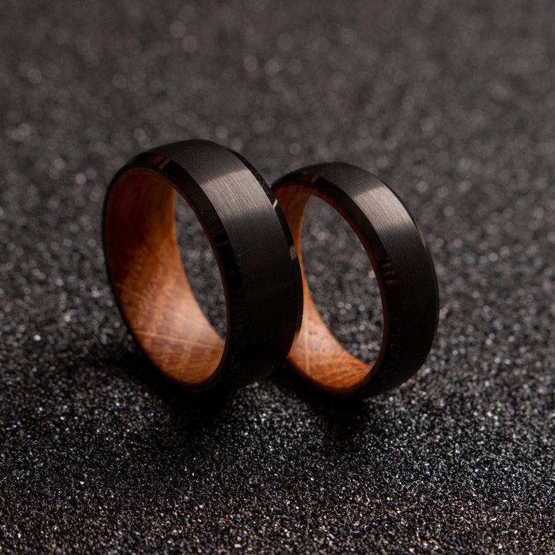 May include: Two black and brown wedding bands. The bands are made of wood and metal. The bands are on a black background.