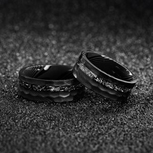 May include: Two black and white rings with textured surfaces and inlaid accents. The rings are set against a dark, speckled background, creating a contrast that highlights their intricate designs. The rings appear to be made of metal.