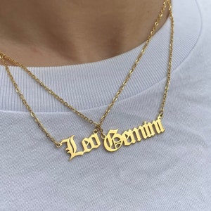 May include: Two gold necklaces with the words "Leo Gemini" in a gothic font.