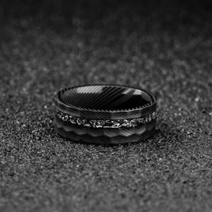 May include: A black and silver ring with a textured band and a decorative silver inlay. The ring has a unique design with a patterned interior and is set against a dark, textured background.