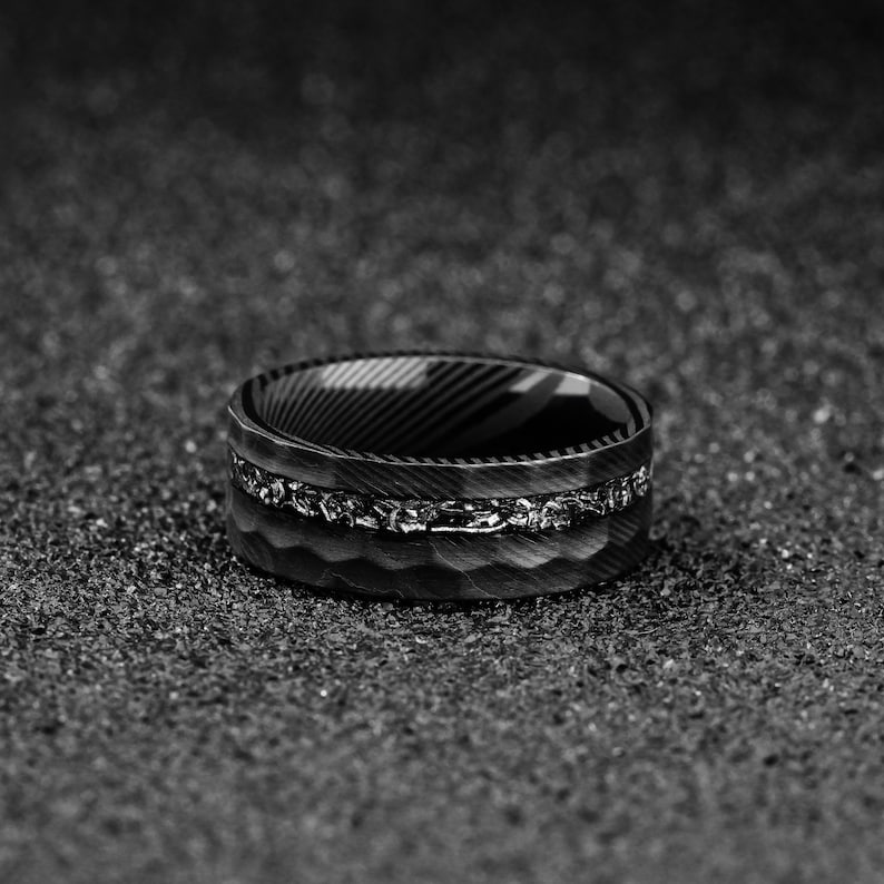May include: A black and silver ring with a textured band and a decorative silver inlay. The ring has a unique design with a patterned interior and is set against a dark, textured background.