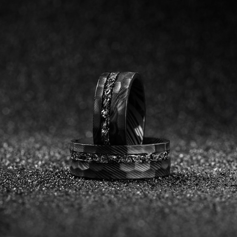 May include: Two black and gray rings with textured surfaces. One ring features a central band of dark stones, while the other has a patterned design. The rings are set against a dark, speckled background.