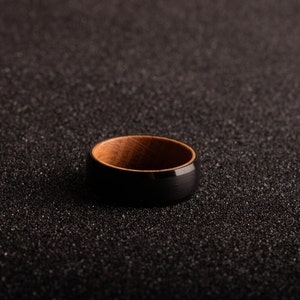 May include: A black and brown ring with a wooden inlay. The ring is on a black textured surface.