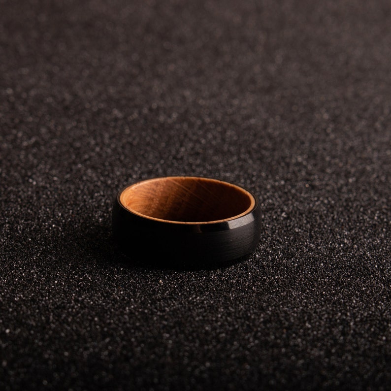 May include: A black and brown ring with a wooden inlay. The ring is on a black textured surface.