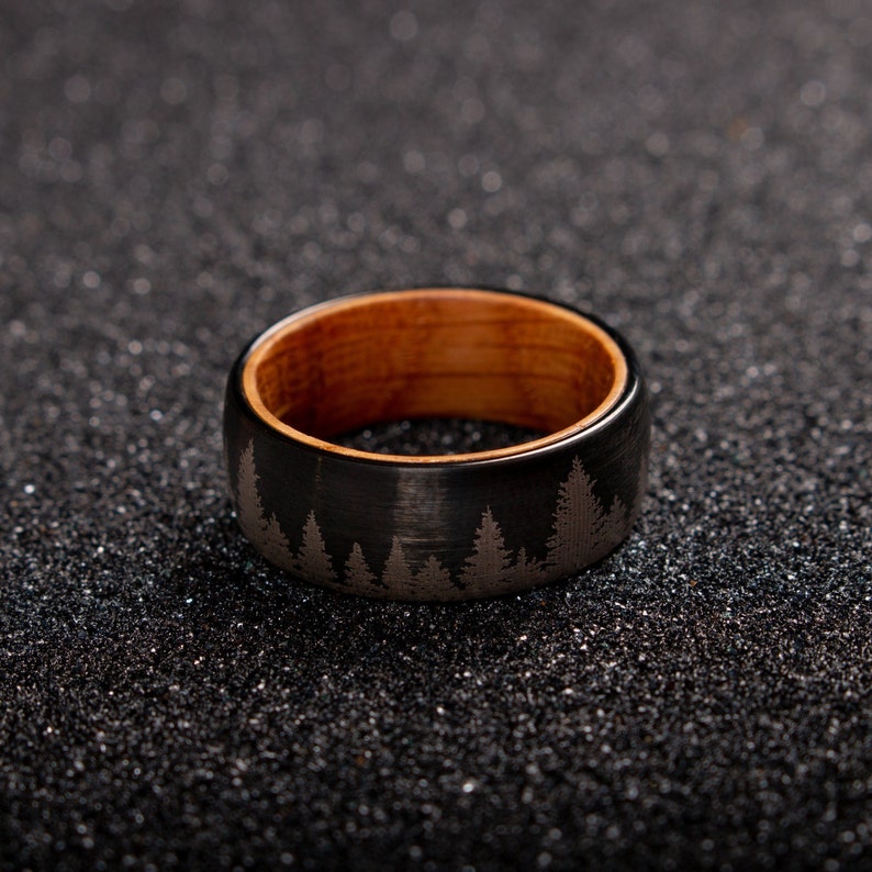May include: A black tungsten carbide ring with a wood inlay and a design of trees etched into the surface.