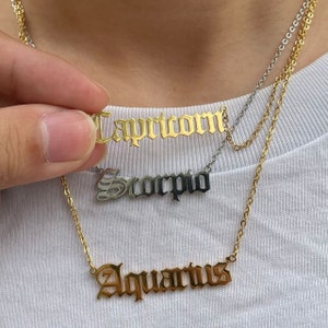 May include: Gold layered necklace with three zodiac signs: Capricorn, Scorpio, and Aquarius. Each sign is written in a gothic font.