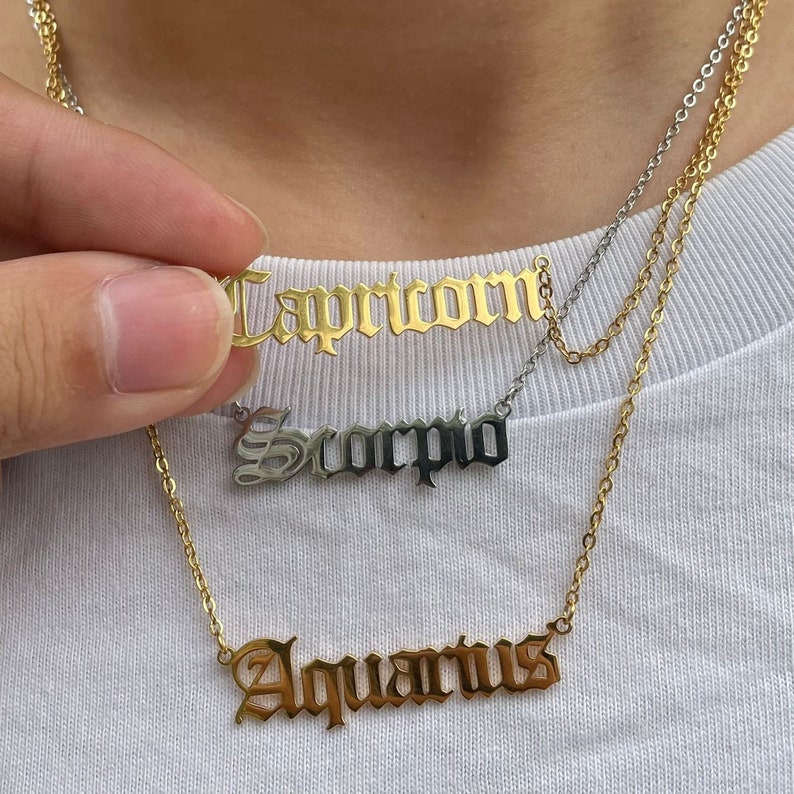 May include: Gold layered necklace with three zodiac signs: Capricorn, Scorpio, and Aquarius. Each sign is written in a gothic font.