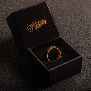May include: A black ring with a gold inner band sits inside a black velvet ring box. The box is closed and has the text "tylian studio" printed in gold on the top.