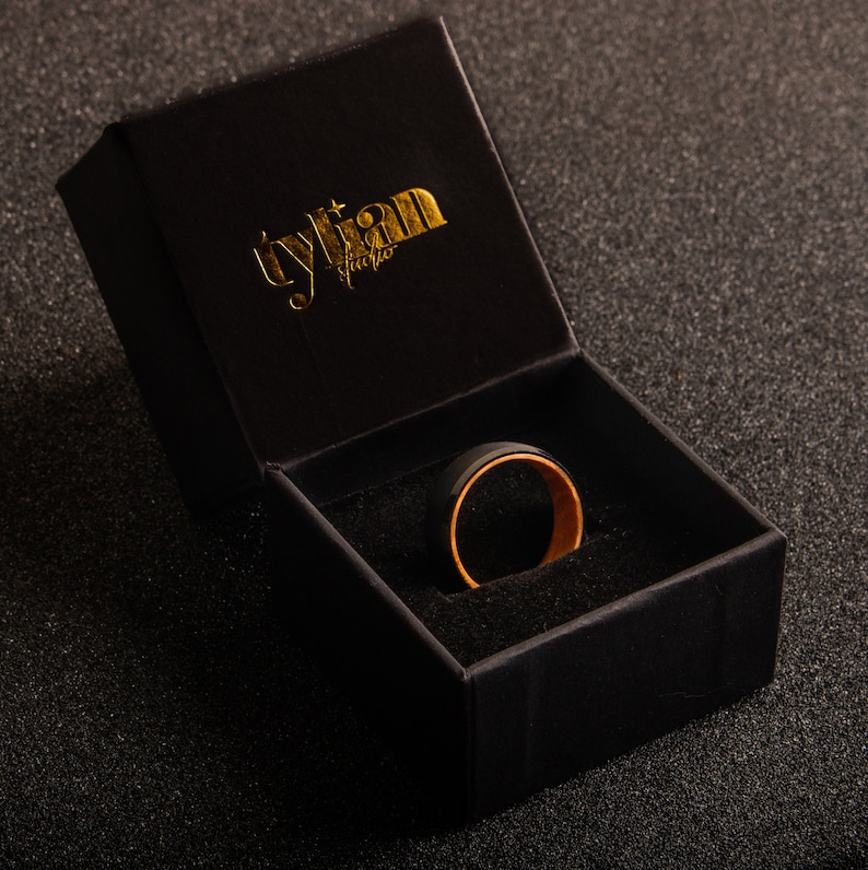 May include: A black ring with a gold inner band sits inside a black velvet ring box. The box is closed and has the text "tylian studio" printed in gold on the top.
