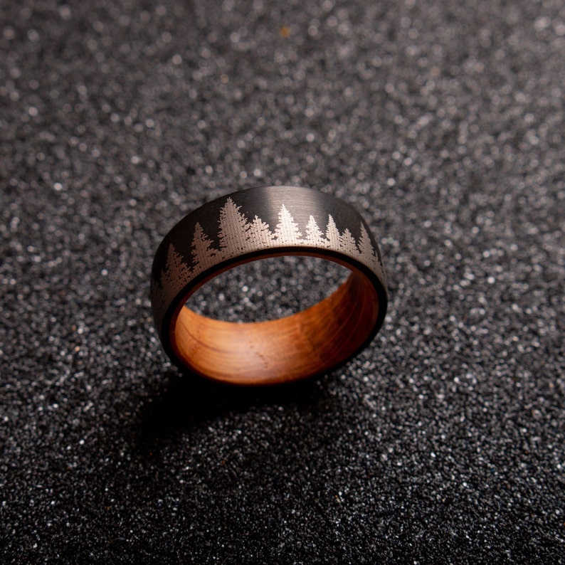 May include: A black tungsten carbide ring with a wood inlay and a silver etched design of a forest.