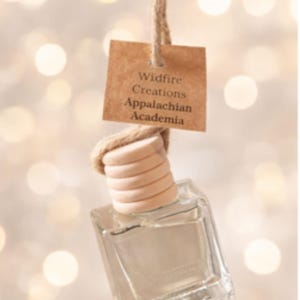 May include: A clear, square glass bottle with a wooden top and a brown tag that reads "Wildfire Creations Appalachian Academia" hangs from a natural twine. The background is a soft, blurred bokeh of warm, glowing lights.