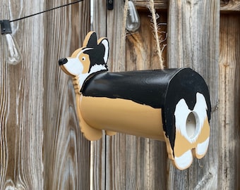May include: A wooden birdhouse shaped like a corgi dog. The birdhouse is painted black, tan, and white. It is hanging from a string.