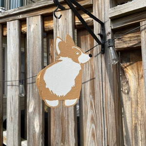 May include: A wooden cutout of a corgi dog hanging from a black metal hook on a wooden fence. The corgi is painted brown and white.