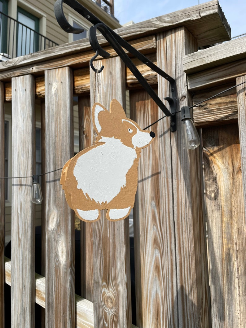 May include: A wooden cutout of a corgi dog hanging from a black metal hook on a wooden fence. The corgi is painted brown and white.