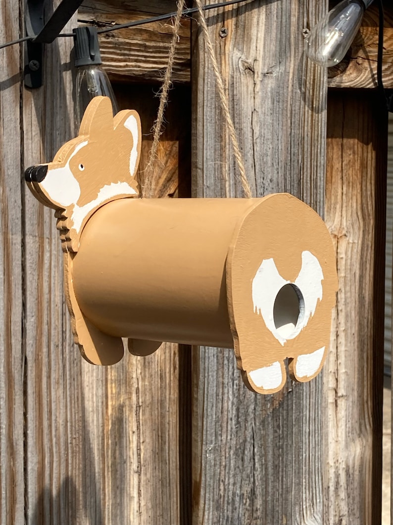 May include: A brown and white wooden birdhouse shaped like a corgi dog. The birdhouse has a round hole for birds to enter and a small opening on the side for cleaning.
