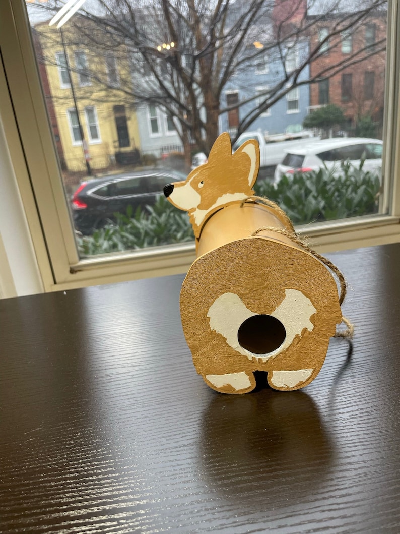 May include: A wooden birdhouse shaped like a corgi dog. The birdhouse is painted brown with white accents. The corgi is looking to the left and has a hole in the back for birds to enter.