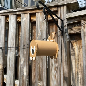 May include: A brown wooden birdhouse shaped like a dog hanging from a black metal hook on a wooden fence. The birdhouse has a white heart-shaped opening and a white tail.