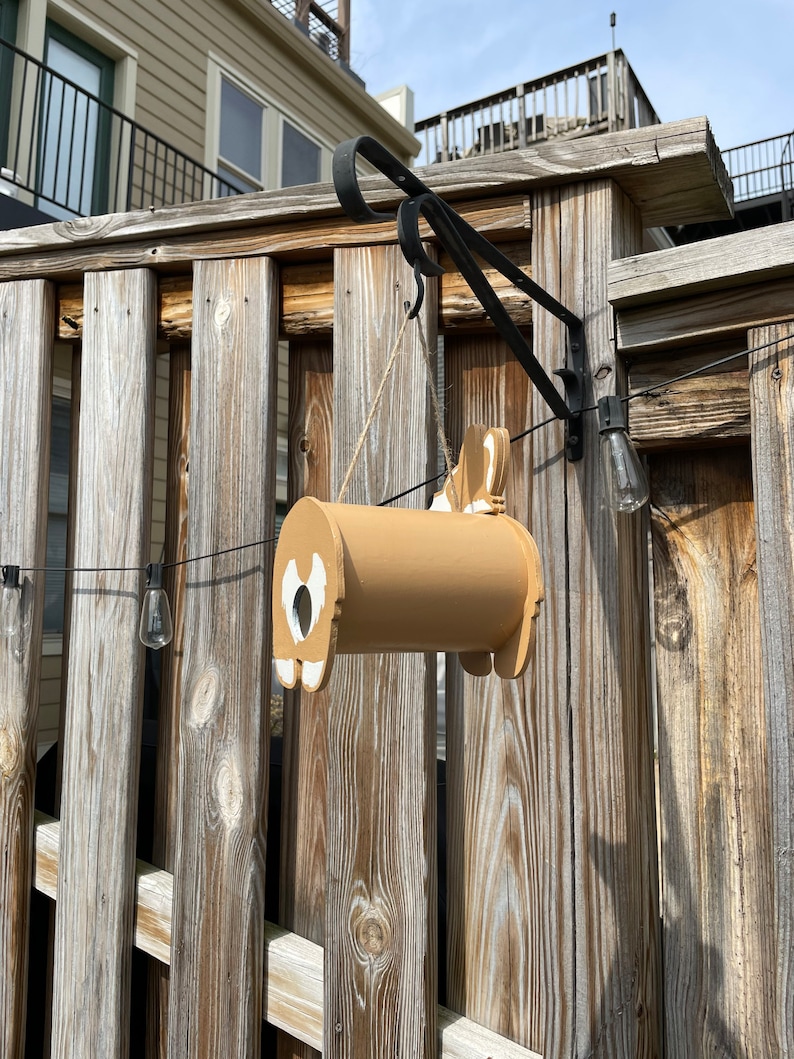 May include: A brown wooden birdhouse shaped like a dog hanging from a black metal hook on a wooden fence. The birdhouse has a white heart-shaped opening and a white tail.