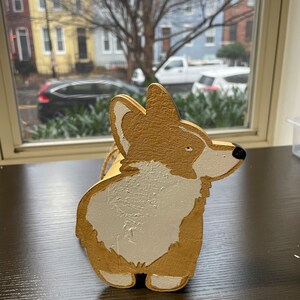 May include: A wooden figurine of a corgi dog painted in gold and white. The dog is standing on its hind legs with its tail curled up.