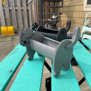 May include: A gray cat-shaped planter made of wood, with a cylindrical container for plants. The cat has a smiling face and a raised tail. The planter is sitting on a turquoise picnic table.