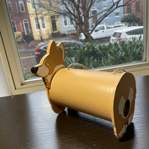 May include: A wooden, hand-painted, brown and white corgi dog shaped birdhouse with a hole in the back for birds to enter.