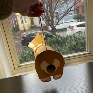 May include: A wooden birdhouse shaped like a corgi dog. The birdhouse is painted brown with white accents and has a hole in the back for birds to enter. The birdhouse is hanging from a string.