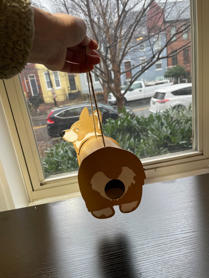 May include: A wooden birdhouse shaped like a corgi dog. The birdhouse is painted brown with white accents and has a hole in the back for birds to enter. The birdhouse is hanging from a string.