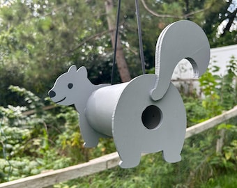 May include: A gray, squirrel-shaped birdhouse hanging from a black cord. The birdhouse is made of wood and features a round body with a tail, head, and feet. The entrance is a circular hole on the back. The birdhouse is suspended in front of a green background.