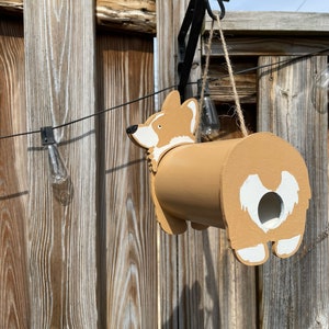 May include: A wooden birdhouse shaped like a corgi dog. The birdhouse is painted brown with white accents and has a small hole for birds to enter. It is hanging from a hook on a wooden fence.