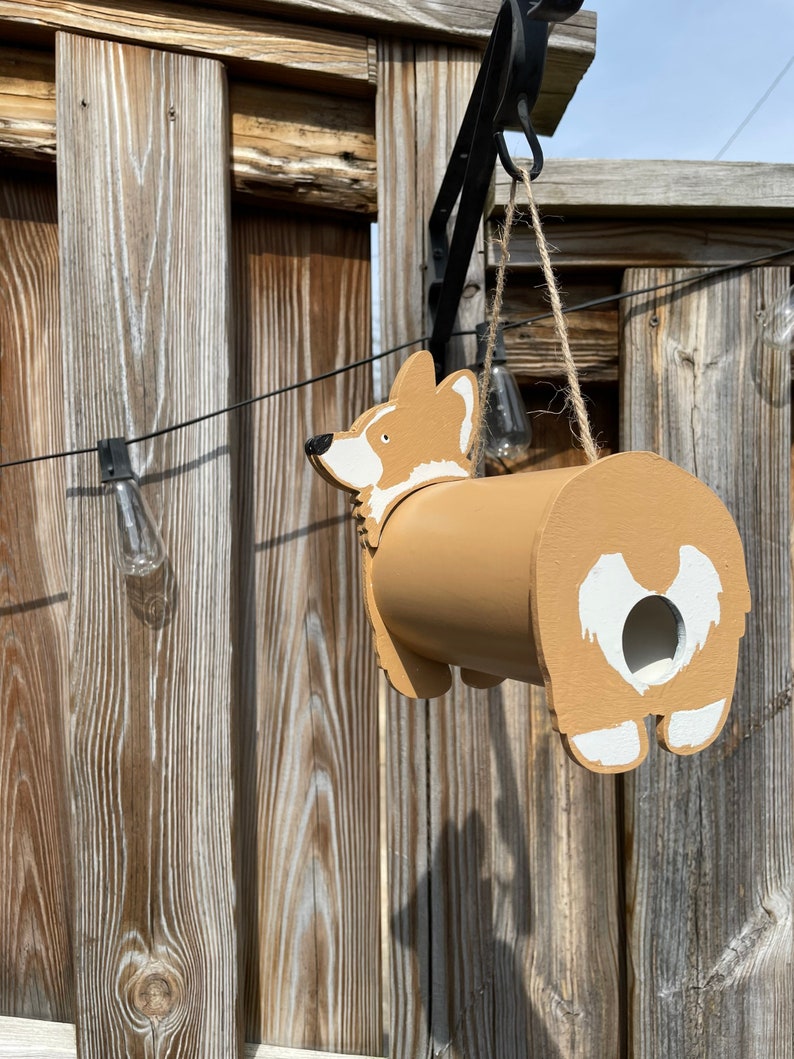 May include: A wooden birdhouse shaped like a corgi dog. The birdhouse is painted brown with white accents and has a small hole for birds to enter. It is hanging from a hook on a wooden fence.