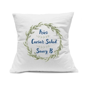 Op de afbeelding: Een vierkant wit sierkussen met een centrale groene botanische krans. Binnenin de krans staat de tekst Fries, Caesar Salad en Sauvy B in een donkerblauw cursief lettertype, gescheiden door kleine horizontale groene takjes als accenten.
