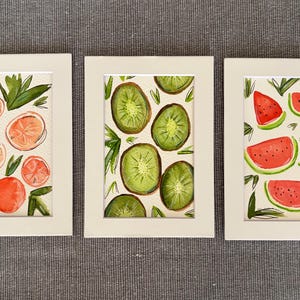 May include: Three framed watercolor paintings featuring fruit. One shows orange slices, another kiwi slices, and the third watermelon slices. Each painting has a white mat and frame, set against a gray background.
