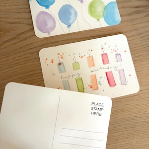 May include: Watercolor birthday cards featuring balloons and candles in various colours. One card reads "happy birthday!". The reverse of a card has lines for writing and the text "PLACE STAMP HERE".