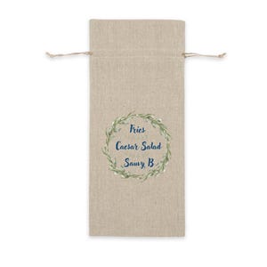 Op de afbeelding: Een rechthoekige beige linnen wijngeschenktas met een trekkoordsluiting aan de bovenkant. Het midden is voorzien van een groen botanisch kransontwerp rondom de donkerblauwe tekst: Fries, Caesar Salad, Sauvy B.