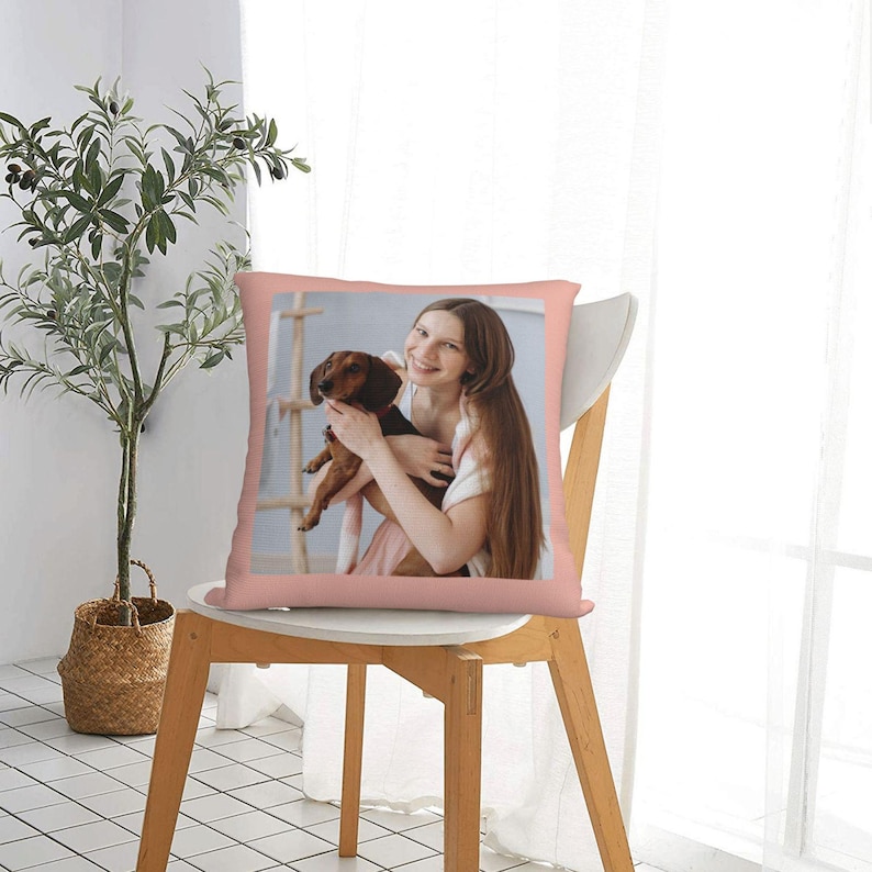 K&ouml;nnte beinhalten: Ein rosa Kissen mit einem Foto einer Frau, die einen braunen und schwarzen Dackel h&auml;lt. Die Frau l&auml;chelt und hat lange braune Haare. Das Kissen liegt auf einem Holzstuhl.