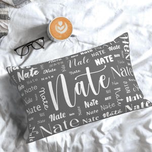May include: A grey rectangular cushion featuring the name "Nate" in white, repeated in different fonts and sizes. The cushion is on a white surface, with a coffee cup and glasses in the background.