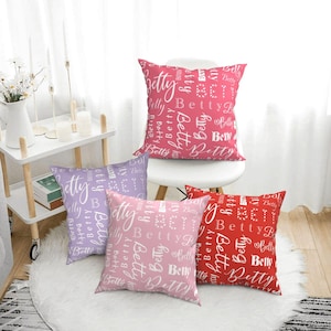 May include: Four decorative throw pillows with a white background and the word "Betty" repeated in various sizes and styles. The pillows are in shades of pink, red, and purple.