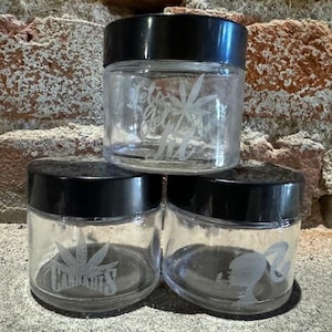 Puede incluir: Tres frascos de vidrio transparente con tapas negras. Los frascos están grabados con diferentes diseños, incluyendo una hoja de cannabis y la palabra "Cannabis".