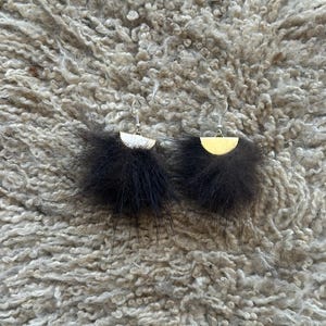 May include: A pair of black feather earrings with a half-moon shaped gold-colored metal base and silver-colored ear hooks. The earrings are displayed on a textured, light gray surface.
