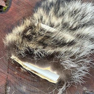 Op de afbeelding: Een close-up van een grijze en witte wasbeerbont met een stuk wit papier eronder. De vacht is zacht en pluche, en de vacht ligt op een houten oppervlak.