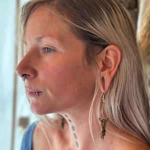 Op de afbeelding: Zijprofiel van een persoon met blond haar die een zilveren kettingoorbel draagt met een bronzen klauw hanger. De oorbel hangt aan een zilveren haak. De persoon heeft meerdere gezichtspiercings.